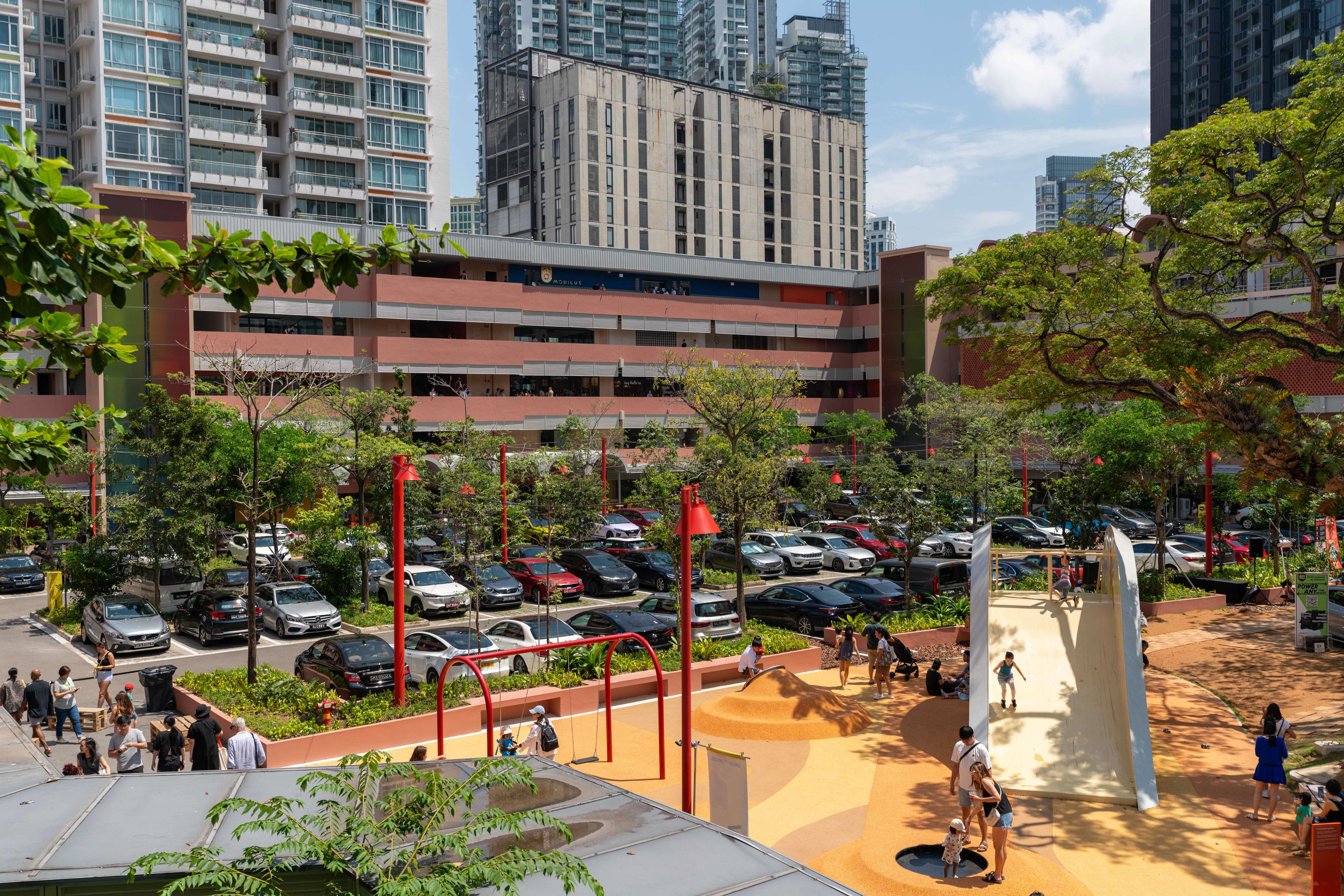 Outdoor scene of a playground area in a city, with a parking lot in the background. Buildings and people are also visible.